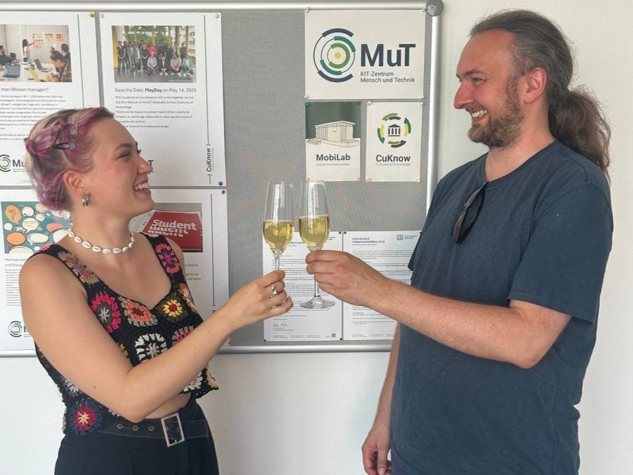 Richard Beecroft (rechts) und Meike Hebich (links) stoßen auf die Übergabe der Geschäftsführung an. Im Hintergrund hängt eine Pinnwand mit dem MuT-Logo.