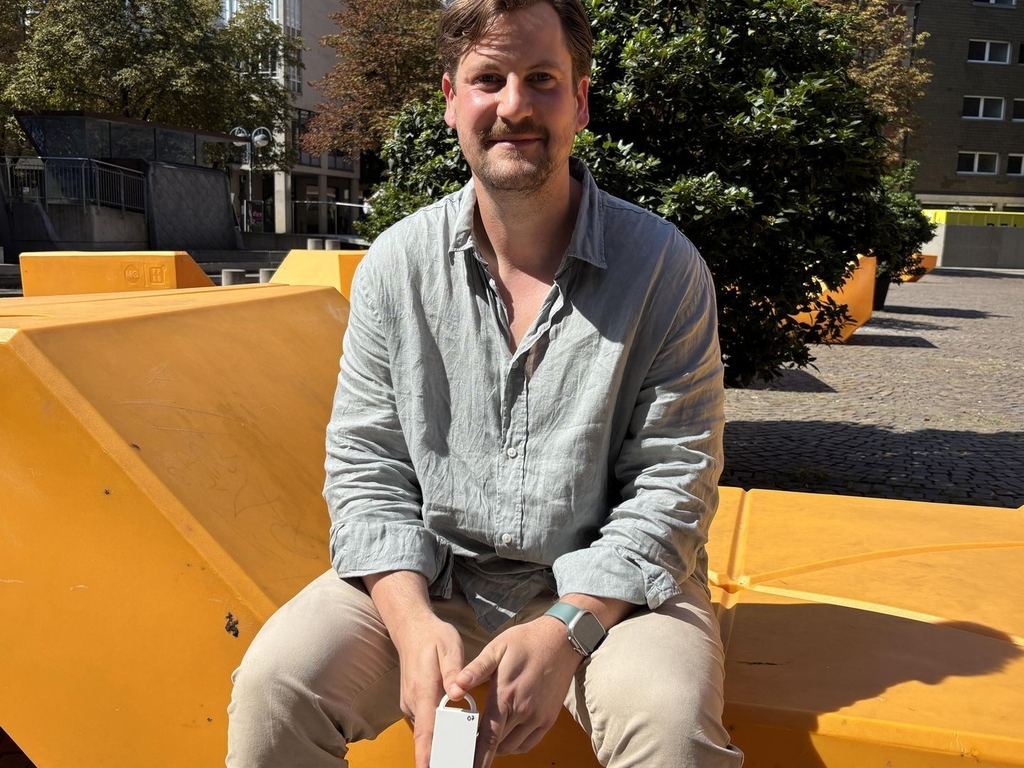 Niklas Heyden sitzt bei Sonnenschein auf orangener Sitzbank im Freien auf dem Kronenplatz, hält ein Smartphone.