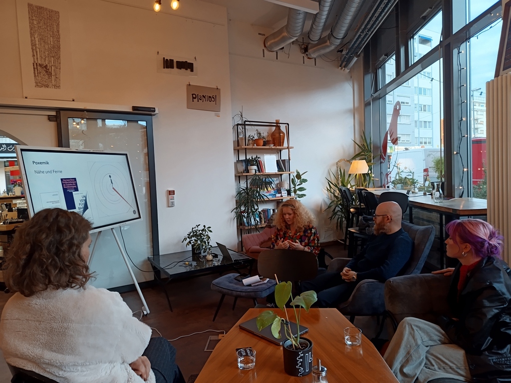 Gruppe in gemütlichem Raum des INTRO Cafés bei Vortrag zu "Nähe und Ferne" an Bildschirm mit Pflanzen.